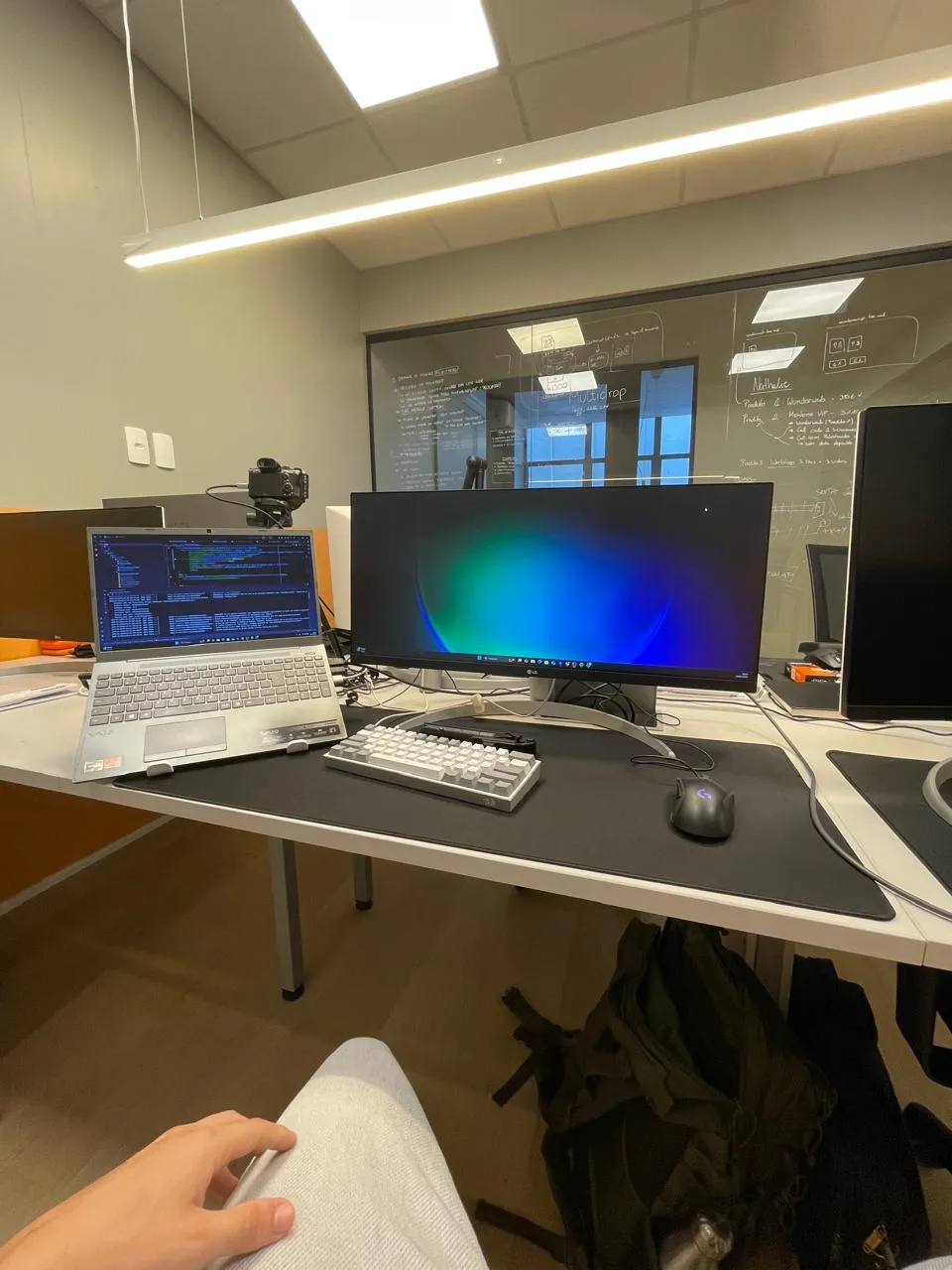 Desk at the Multidrop office in Florianópolis, with an ultrawide monitor and a glass wall covered in notes