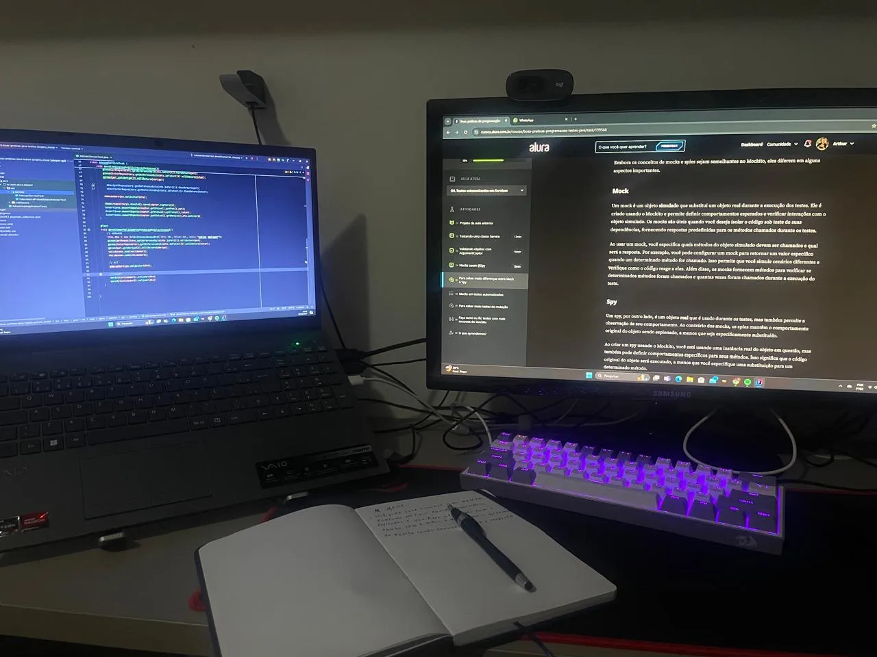 Desk with a laptop showing Java code, a monitor with an Alura course and a notebook
